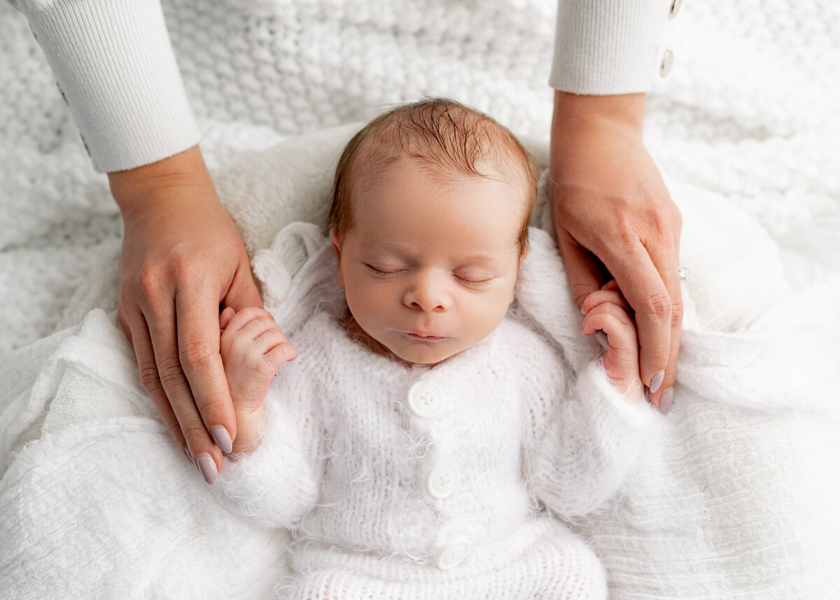 mom holding sleeping baby's hands on a white blanket. baby is wearing a white knit outfit