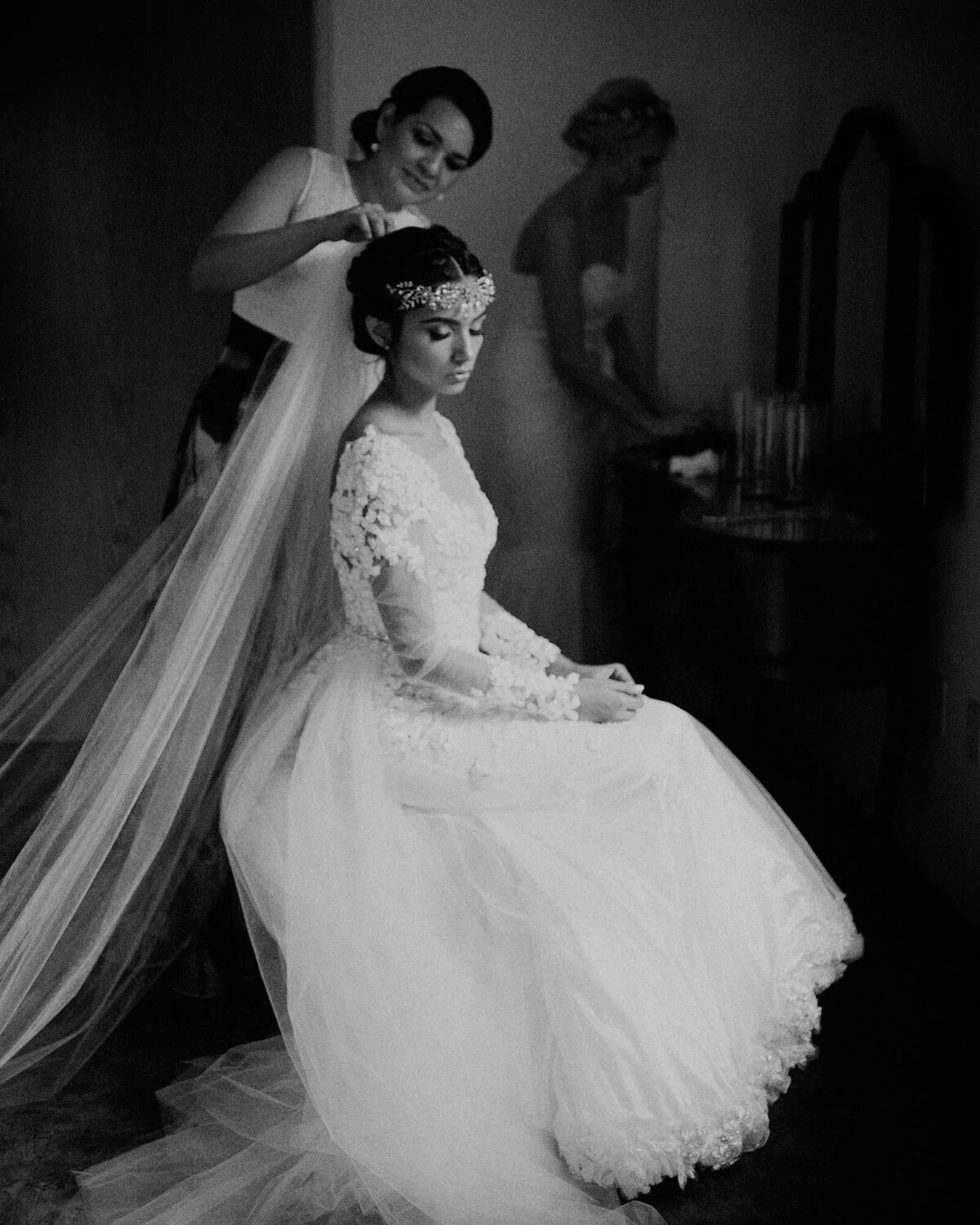 A black and white photograph of a bride seated while another person adjusts her veil