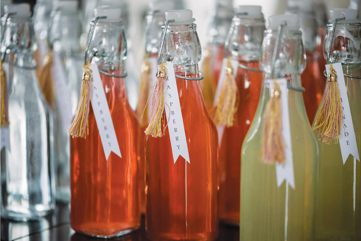 Array of individual bottled drinks with custom labels and pink and yellow tassels, set up for a girl’s baby shower.