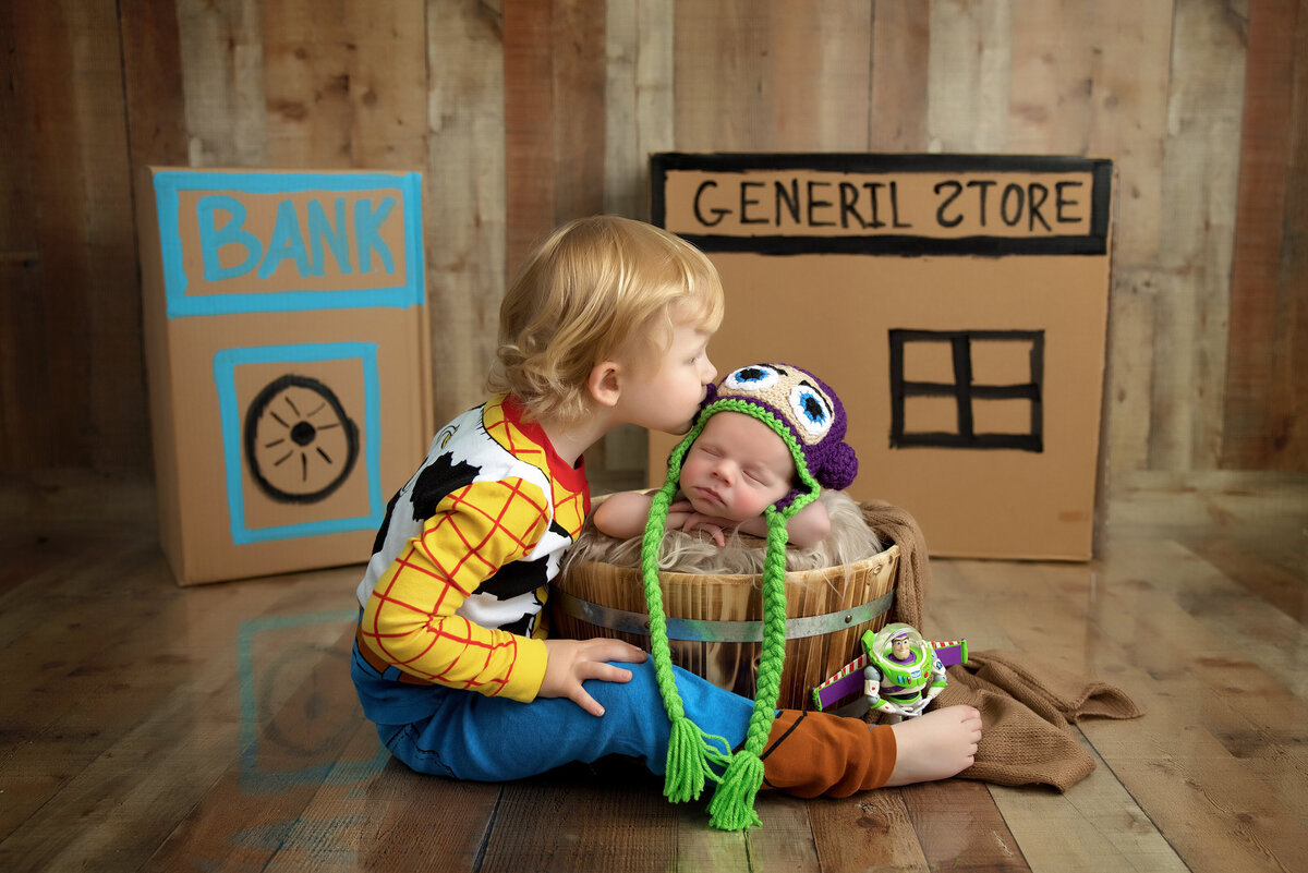 A child dressed as Woody from Toy Story tenderly kisses a sleeping baby in a colorful crocheted hat nestled in a basket. The scene, expertly captured by a Jacksonville baby photographer, features cardboard cutouts resembling a bank and general store.