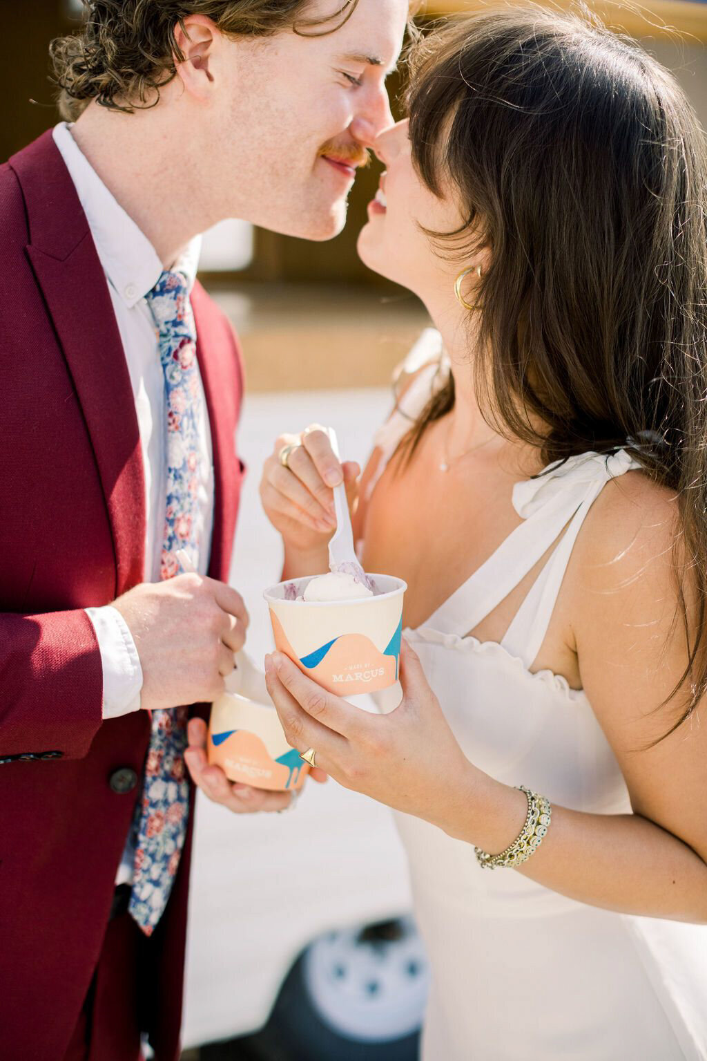 Couple eating ice cream from Groom wearing burgundy suit and floral tie, bride in sleek modern bridal gown in front of Made By Marcus, unique and playful ice cream based in Calgary, AB. Featured on the Brontë Bride Vendor Guide.