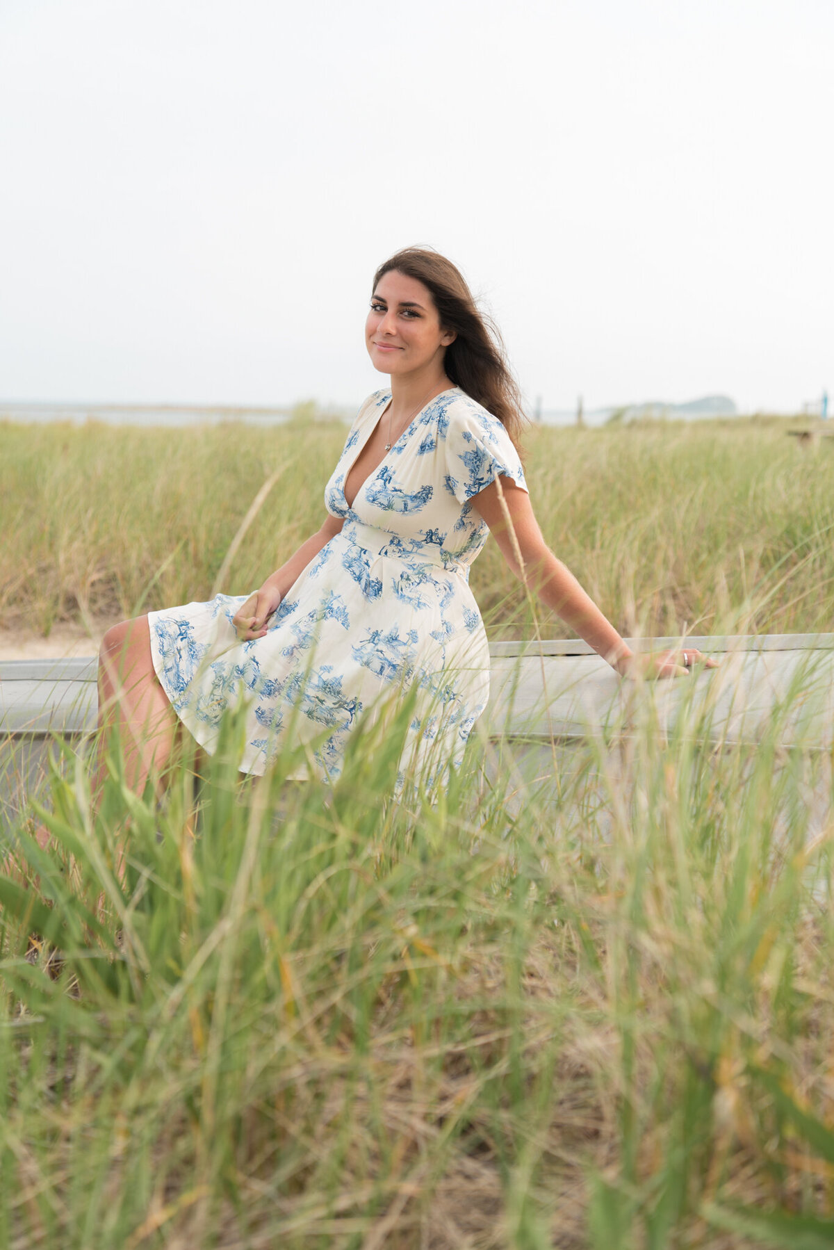 Teen girl sitting on a piece of driftwood in dune grass with white dress, blue flowers.