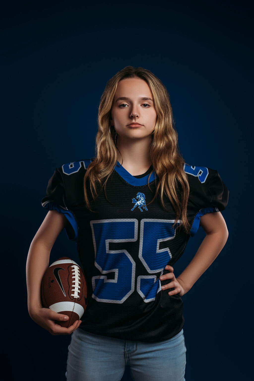 A teenage girl in a sports jersey holds a football in front of a blue solid backdrop for her senior graduation photoshoot, captured by a fine art studio photographer in Bemidji, Minnesota