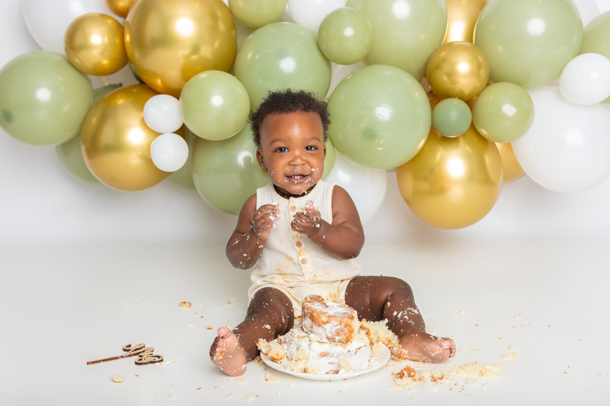 Baby eating a simple white cake with an olive and gold balloon backdrop