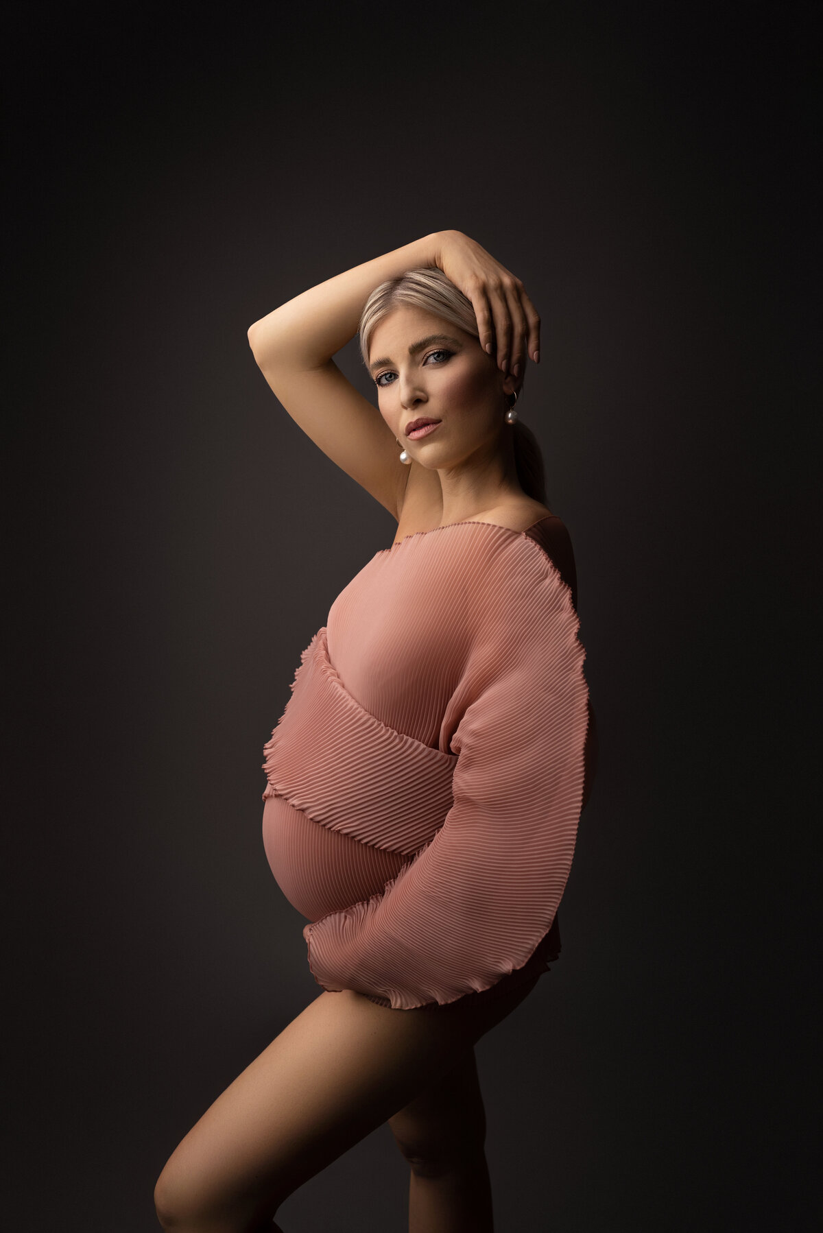Woman poses for a fine art maternity photoshoot with Philadelphia's best maternity photographer Katie Marshall. She is standing side profile to the camera with her foreleg bent. Her forearm is underneath her baby bump, her back hand is resting gently atop of her head. She is looking over to ward the camera with a serious expression.