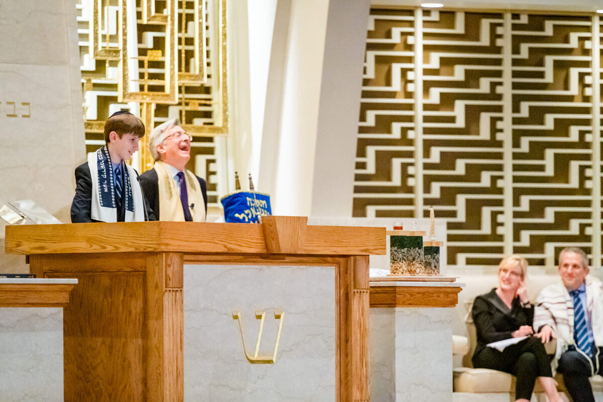 A rabbi laughs while a boy stands at the bimah reading the torah
