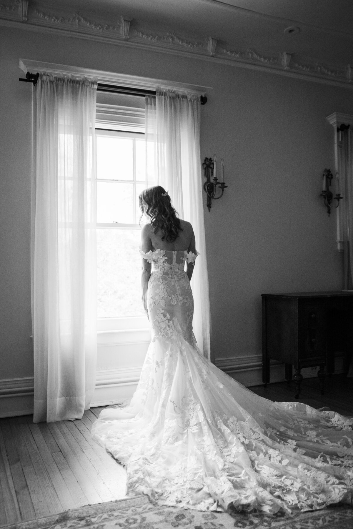 A bride faces away from the camera in her wedding dress as she stares out the window.