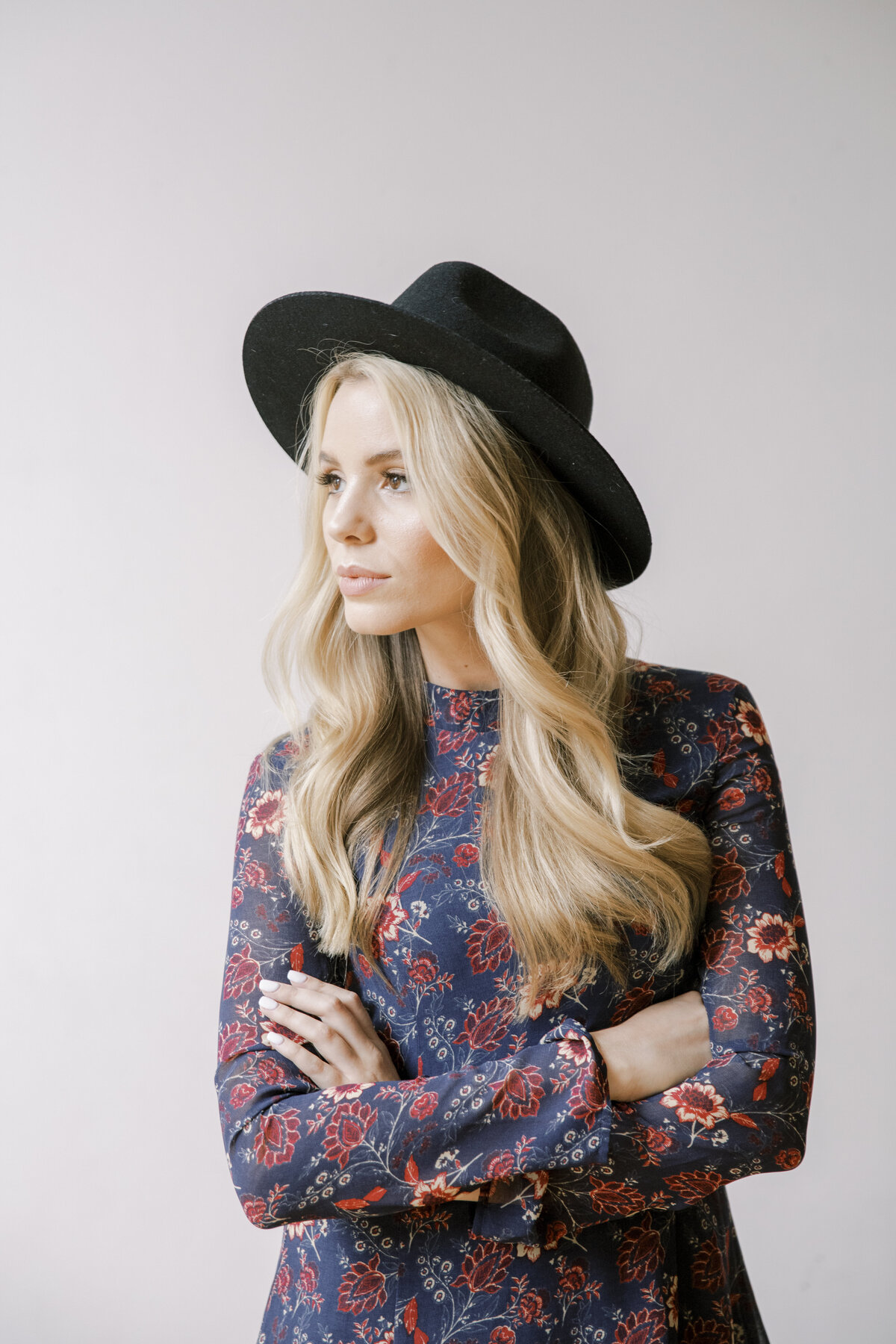 blond woman with black hat and floral top crossing her arms in front of her and looking off into the distance
