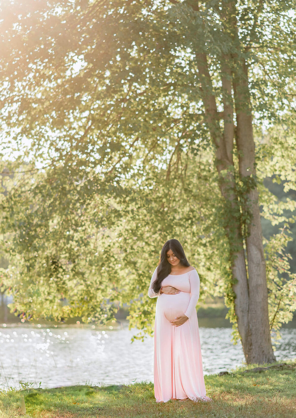 gorgeous mom to be standing near a tree on a sunny summer day, the wind moves her hair and dress, she is smiling and looking down at her belly