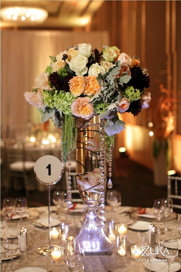 elevated arrangement of peach roses, ivory roses, green hydrangea, dusty miller, green amaranthus, on top of crystal glass vase with crystal bead strands