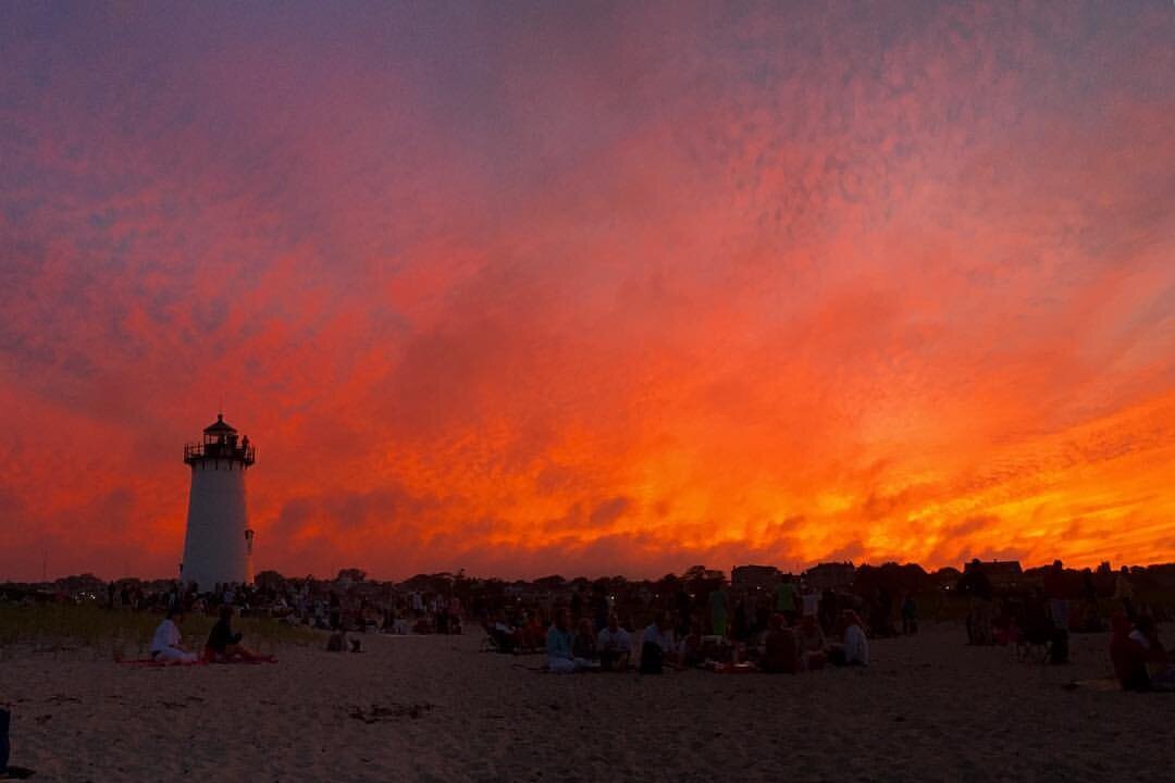 photo of a brilliant sunset sky at Martha's vineyard; the silhouette of a white lighthouse punctuates the sunset, and the beach at dusk is peppered with people