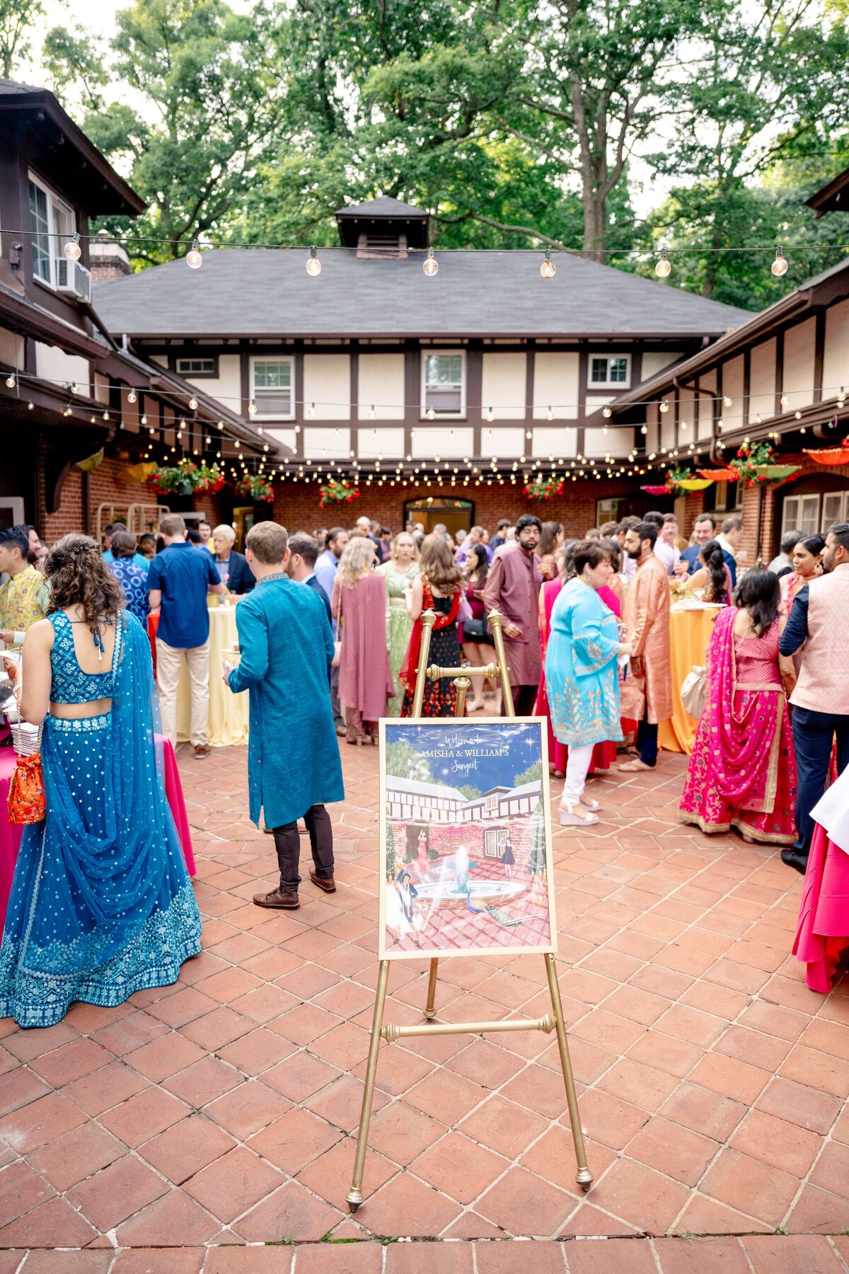 A vibrant outdoor gathering with people dressed in colorful traditional attire. The scene features a courtyard with string lights, surrounded by buildings. A decorative easel with a picture stands in the foreground.