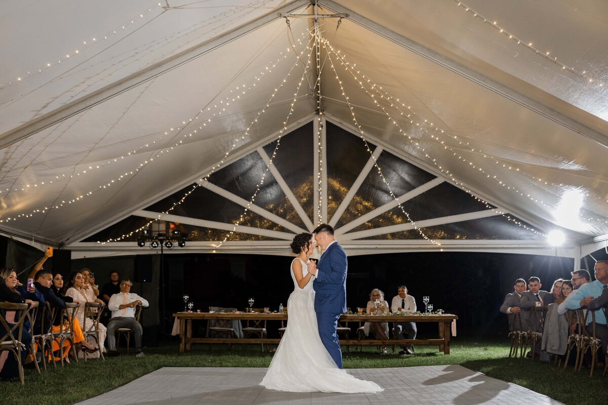 A bride and groom share their first dance under a tent adorned with string lights, with guests watching and clapping around them at an event in Davenport.