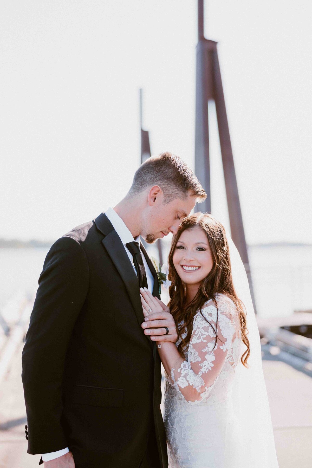 A groom nuzzles his bride while standing on a dock as she smiles with a hand on his chest