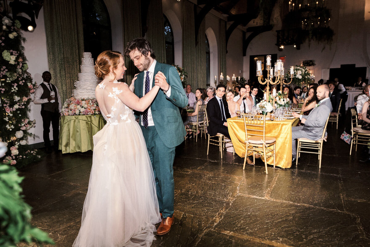 The bride and the groom are dancing while guests, sitting on dining tables, watch them.