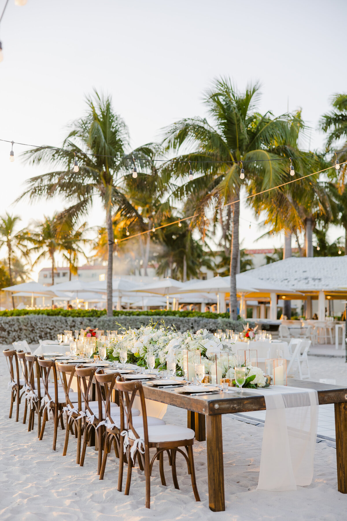 Outdoor dining setup on a beach at sunset with wooden tables, floral decorations, and string lights amidst palm trees, captured by a Destination Wedding Photographer.