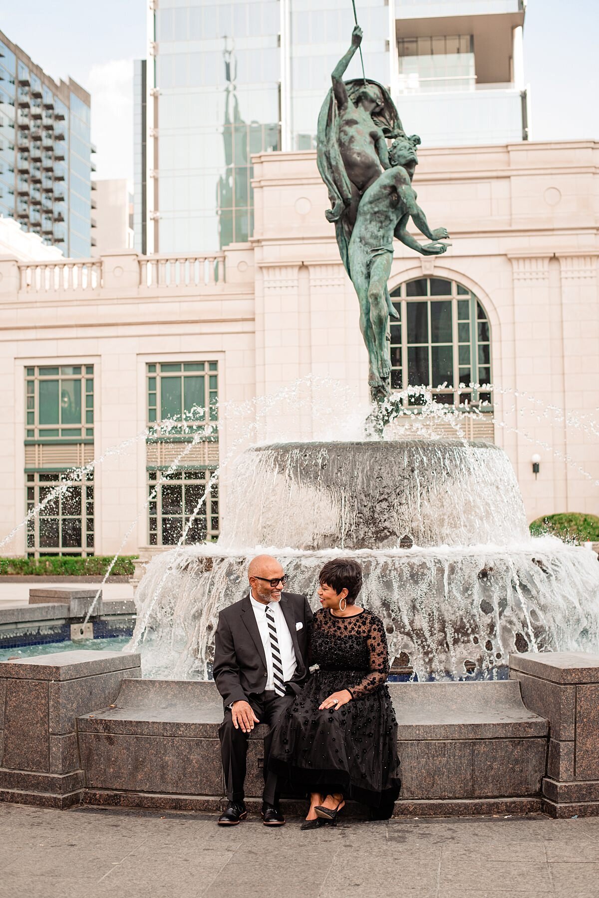 The bride and groom sit on the edge of the fountain at the schermerhorn symphony center in Nashville. The groom is wearing a black suti with a white shirt with a black and white striped tie. The bride is wearing a black dress with a sheer neckline and sheer long sleeves. The dress is adorned with tiny black flowers with rhinestones in the center of each flower.
