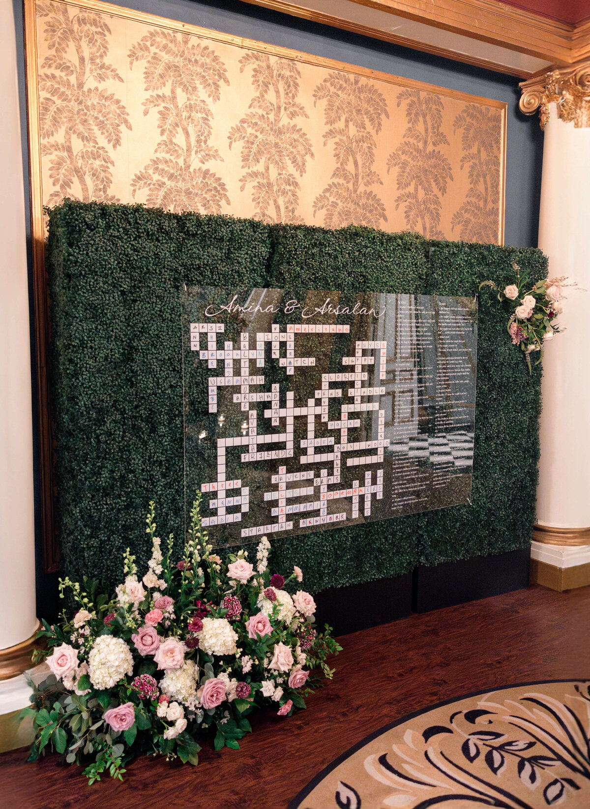 An elegantly decorated wall display at a wedding features a large crossword puzzle with green foliage backing and pink floral arrangements at the base. Names are written above the puzzle in cursive.