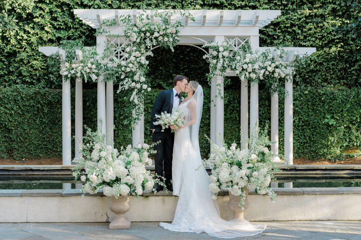 William Aiken House arbor decorated in white and green flowers with bride and groom kissing. Pedestal flowers. Spring wedding in Charleston. Destination wedding photographer.