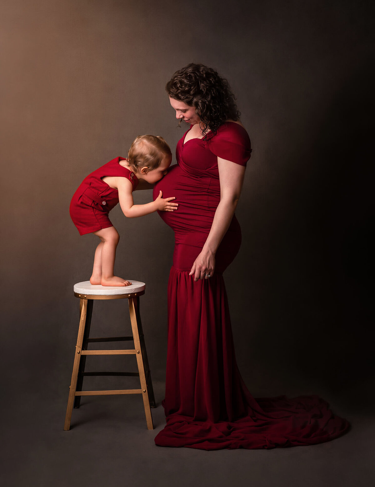 An Expecting mama in a long red dress  stands still as her son kisses her baby bump while standing on a stool