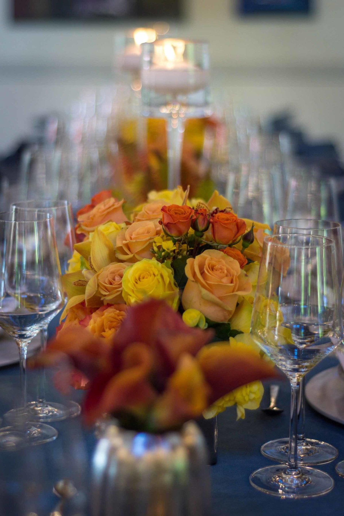 lont table lined with centerpieces of orange roses and orange calla lilies  on blue linen