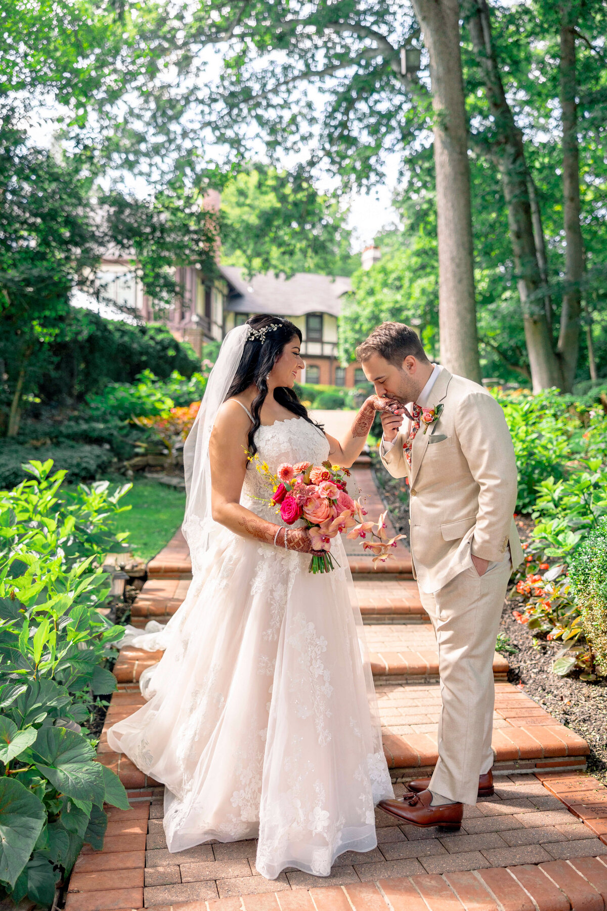 A bride and groom share a tender moment on a garden path. The bride, in a lace gown, holds a vibrant bouquet. The groom, in a beige suit, gently kisses her hand. Lush greenery and a charming house provide a scenic backdrop.