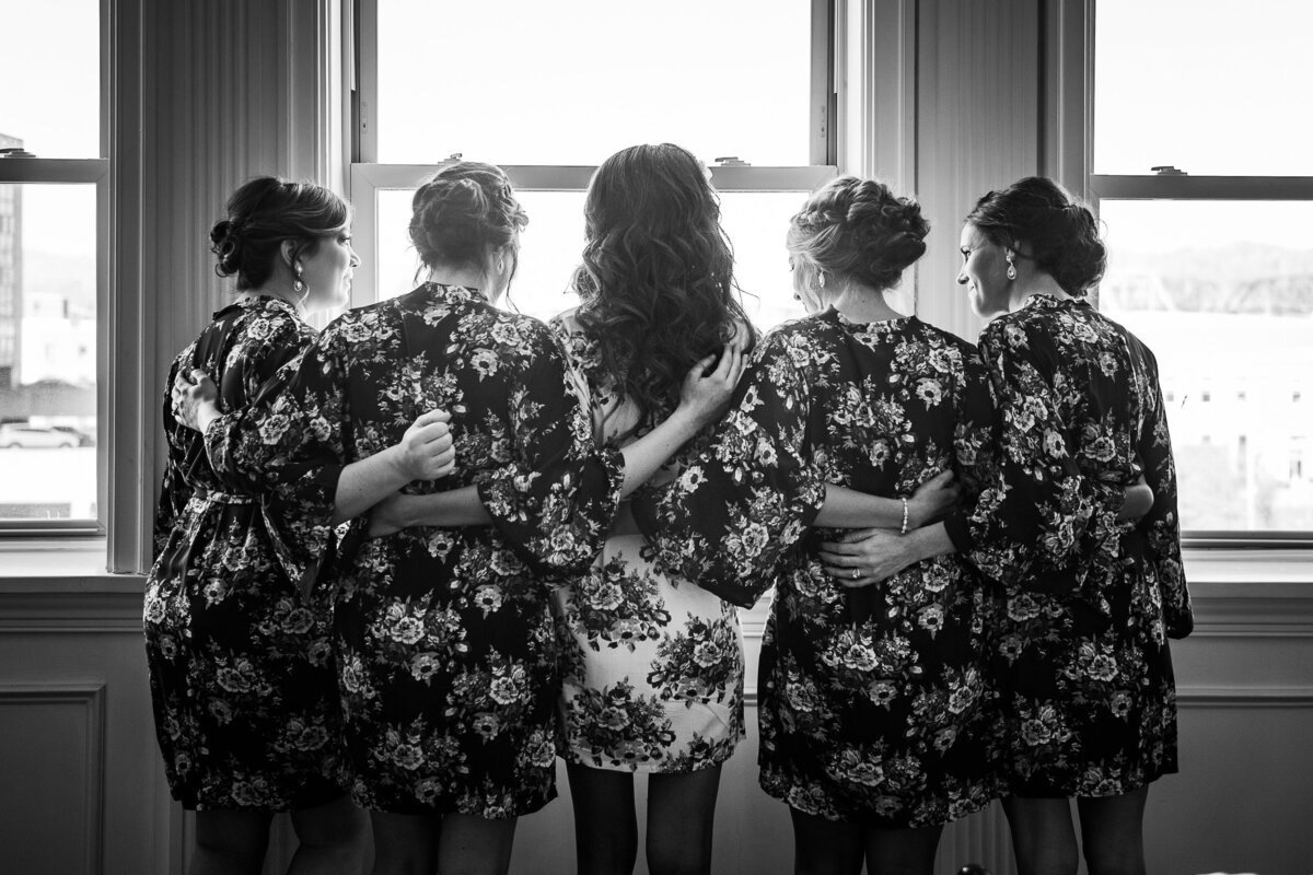 black and white image of bride with bridesmaids arms linked looking out window