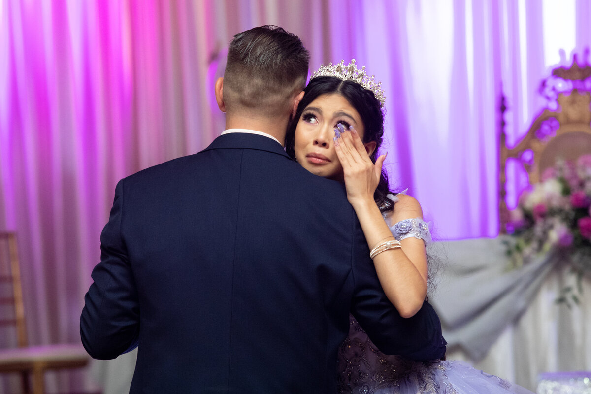 A teenage girl in a purple ballgown wipes away tears while dancing with her father in a dark blue suit New York Westchester quinceanera Photographer Milestone images