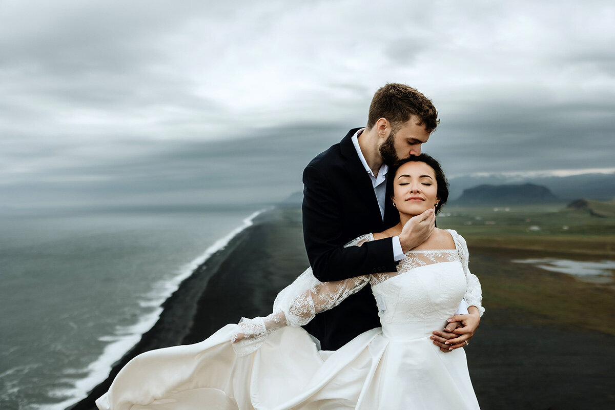 groom is kissing his bride on her head black sand beach and ocean in the background they are in Iceland