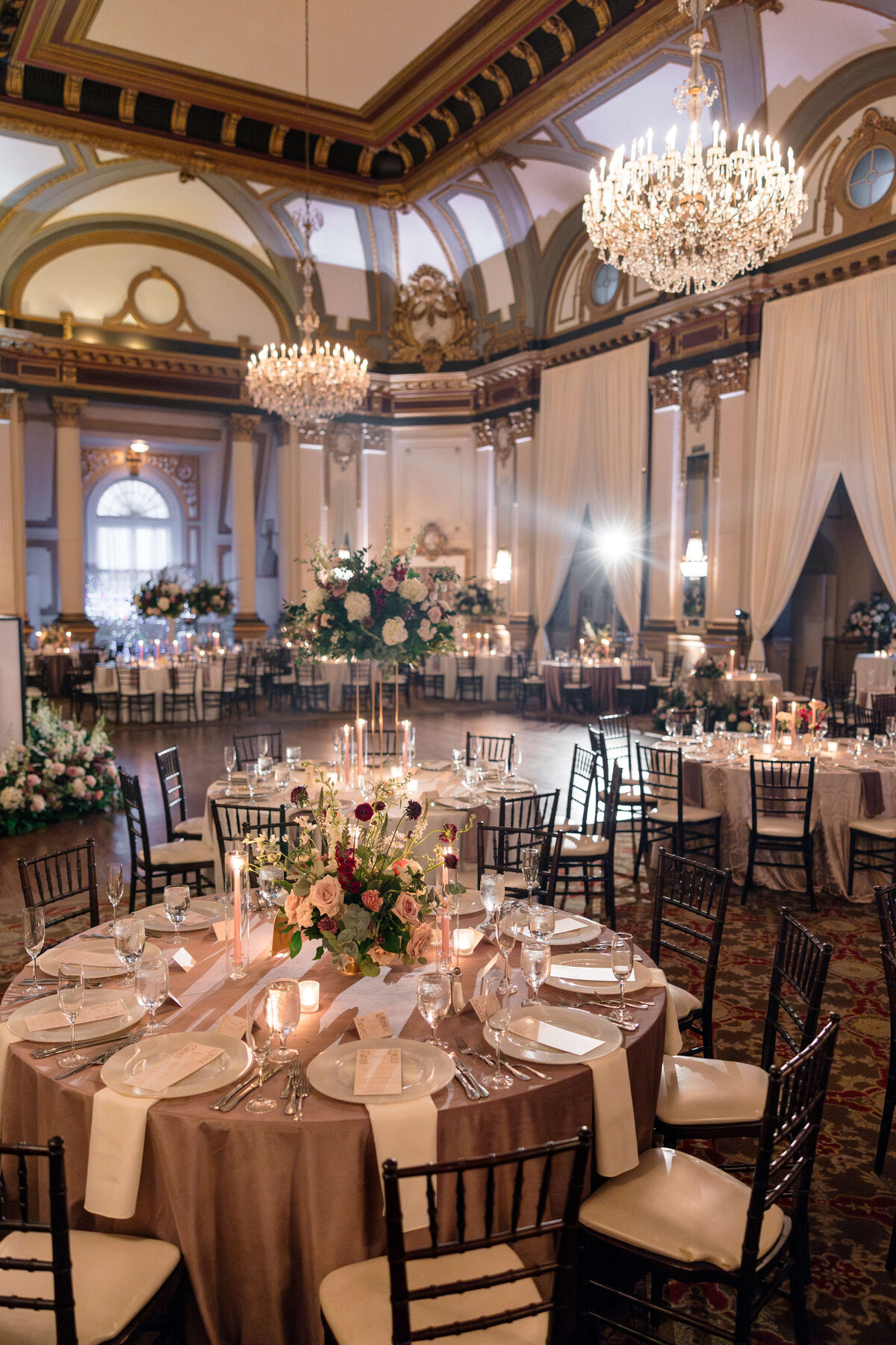 A lavishly decorated ballroom set for a formal event. Round tables are adorned with elegant centerpieces of flowers and candles. The room features high ceilings, large chandeliers, and draped windows, creating a sophisticated atmosphere.