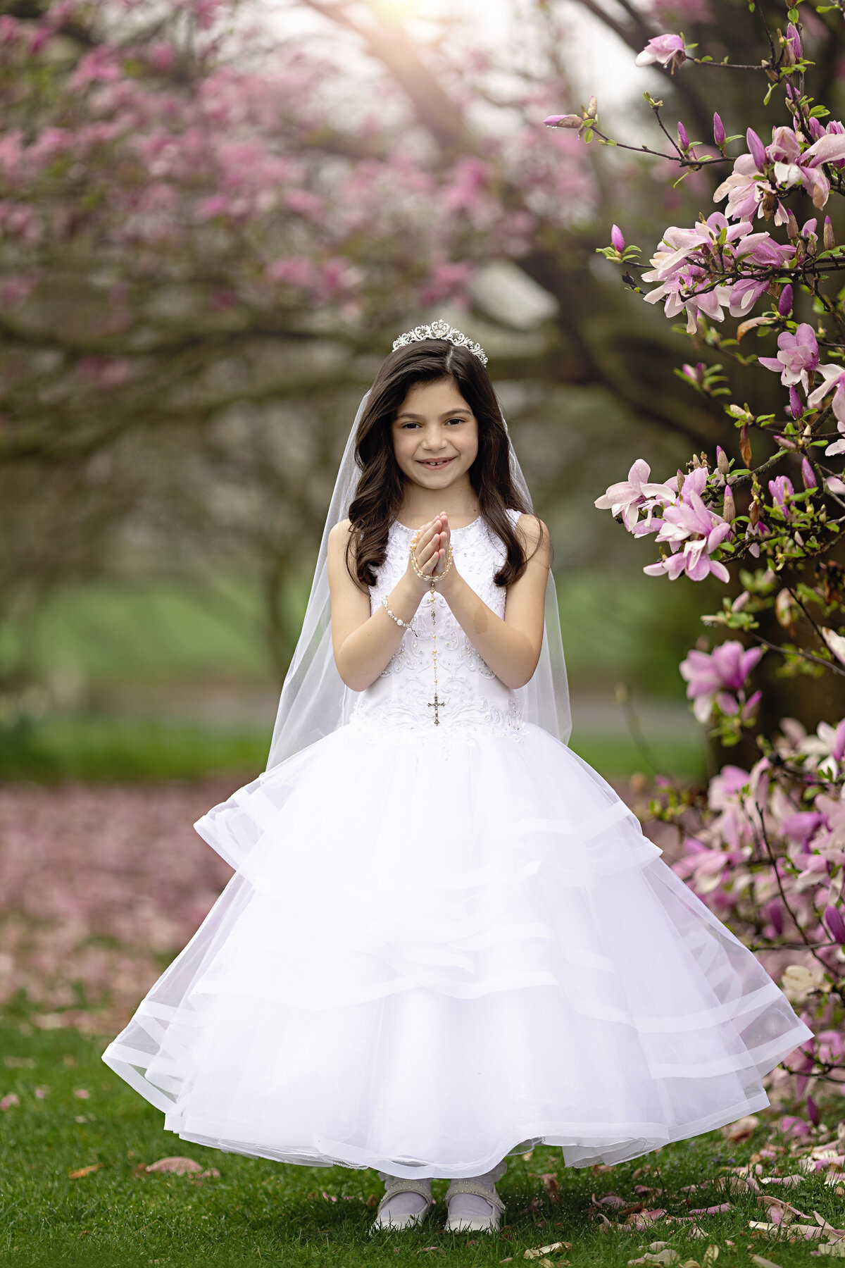 A girl in a white dress prays with some rosary beads before her communion among some pink flowering trees