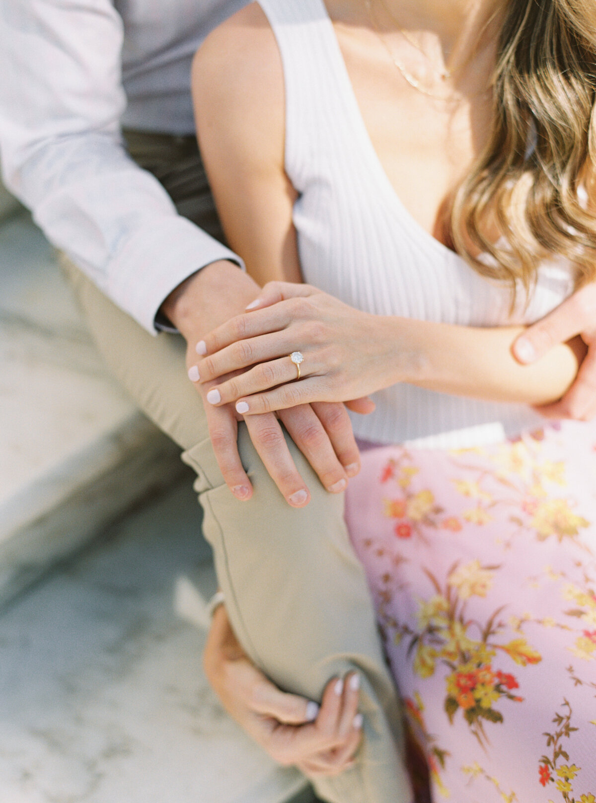 Pink floral skirt. Girl sitting in between legs of guy with engagement ring holding hands. Downtown Charleston spring engagement session. Kailee DiMeglio Photography.