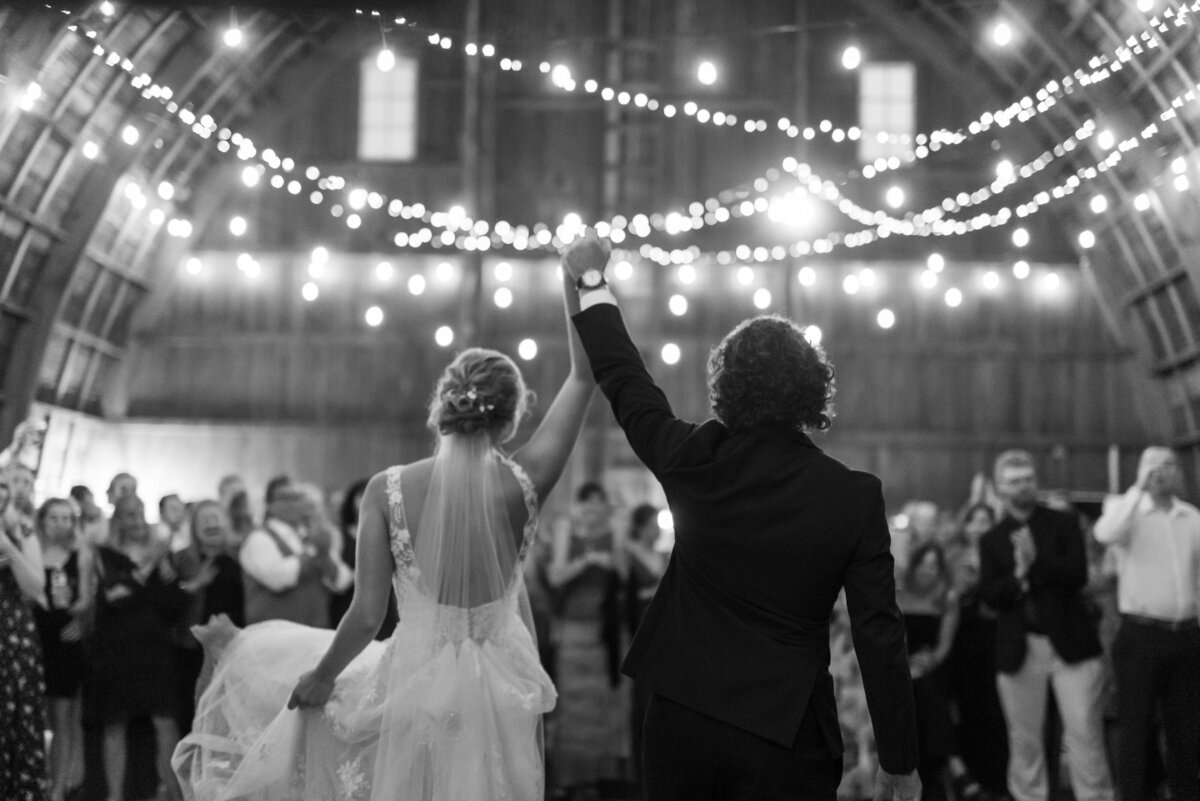 Bride and groom walk into their string light lit reception and raise their hands in the air in celebration as their guests cheer