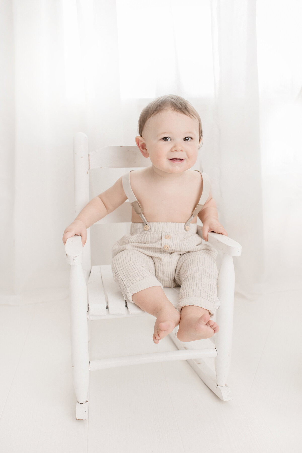 one year old boy in suspenders sitting in white rocking chair