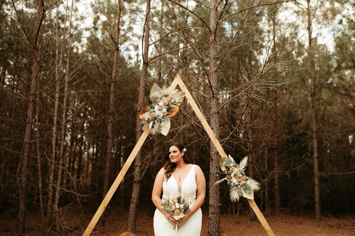 Bride holding dried floral bouquet standing underneath the triangle arch in front of trees