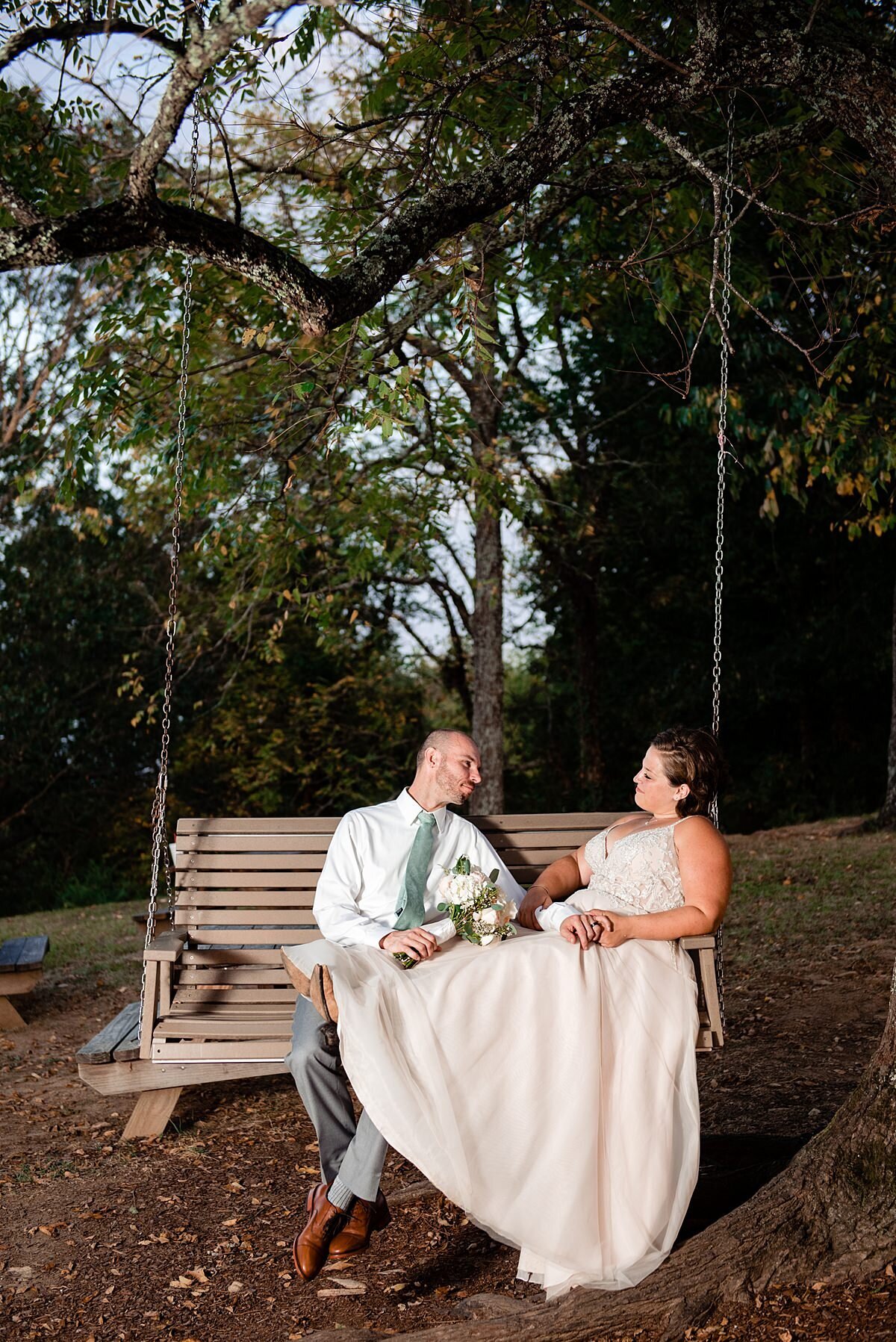 The groom sits on a wooden swing suspended from a tree as he slowly rocks the bride back and forth. The bride sits with her feet up on the groom's lap with her back to the side of the swing her heavy satin wedding dress spilling down to the ground. The brides dress is sleeveless with a fitted bodice. The groom is wearing gray pants with a white shirt and a light gray tie.