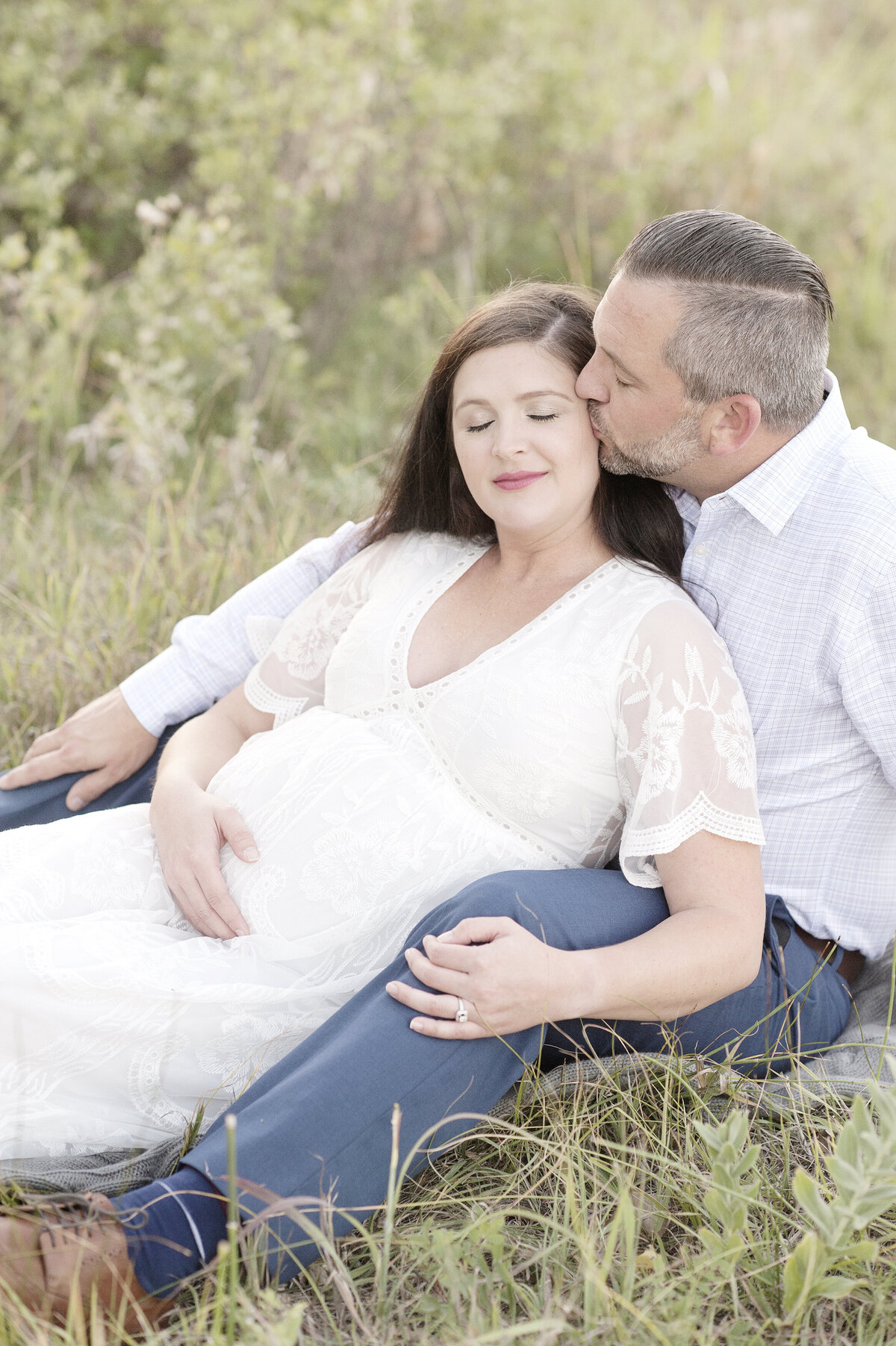 Man kissing pregnant wife in white dress sitting in tall grass