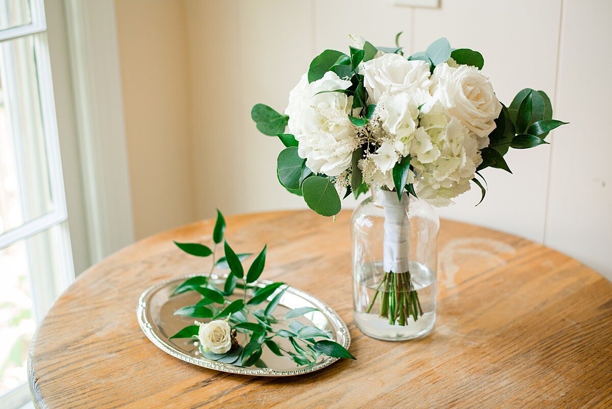 The bride's white bouquet sits in a glass vase on a wooden table beside a small silver tray with the groom's white boutonniere accented with greenery.