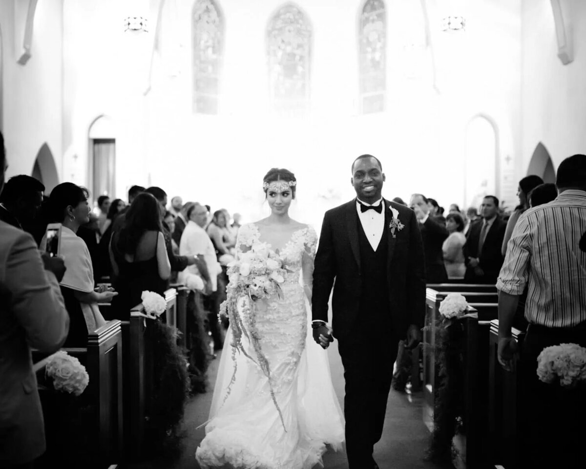 A bride and groom walking down the aisle, post-ceremony, surrounded by guests in a classic black and white photo