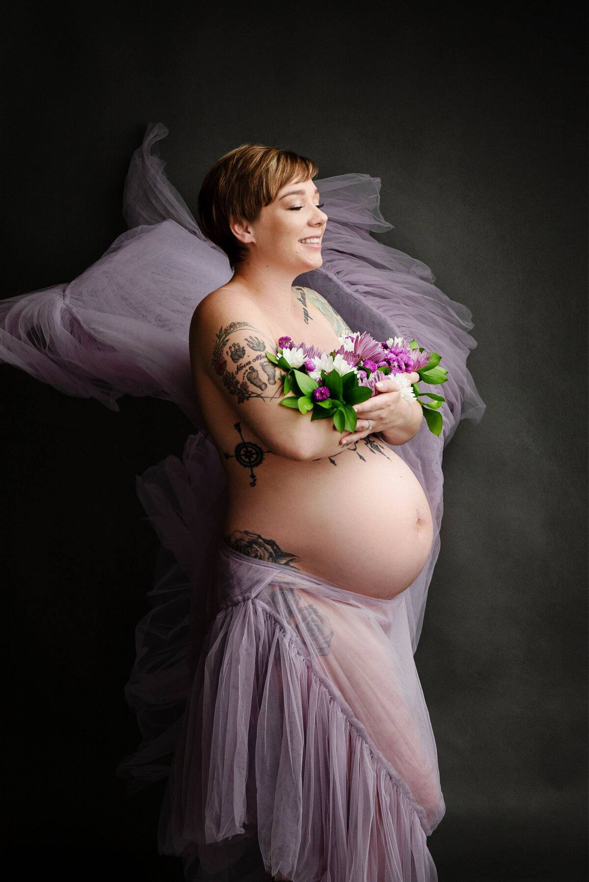 st-louis-maternity-photographer-expecting-mother-nude-with-purple-fabric-covering-her-while-she-cradles-a-bouquet-of-flowers