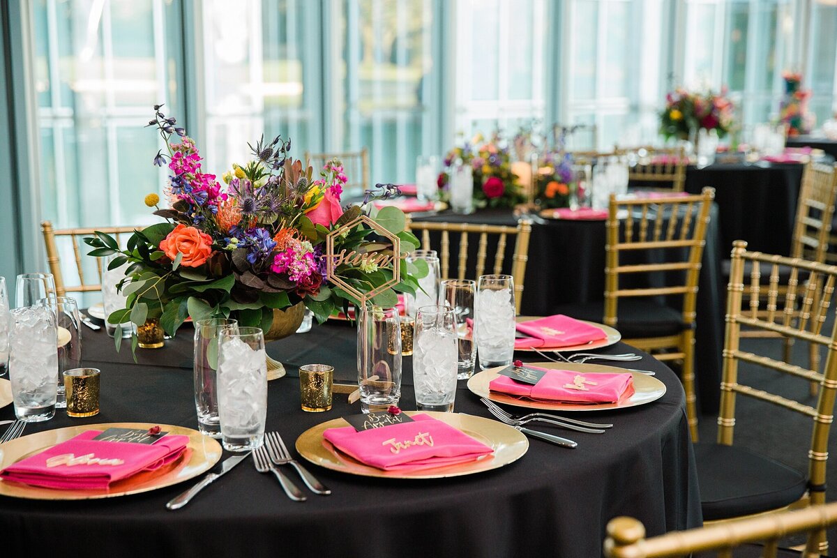A detail shot of a reception table at a wedding at the Modern Art Museum of Fort Worth in Fort Worth, Texas. The large round table is covered by a black tablecloth, gold plates, pink napkins that have the names of the guests on them in gold, cutlery, glasses, candles, and a centerpiece of colorful flowers. The table is surrounded by gold chairs with black cushions. Similar tables and chairs can be seen in the background.