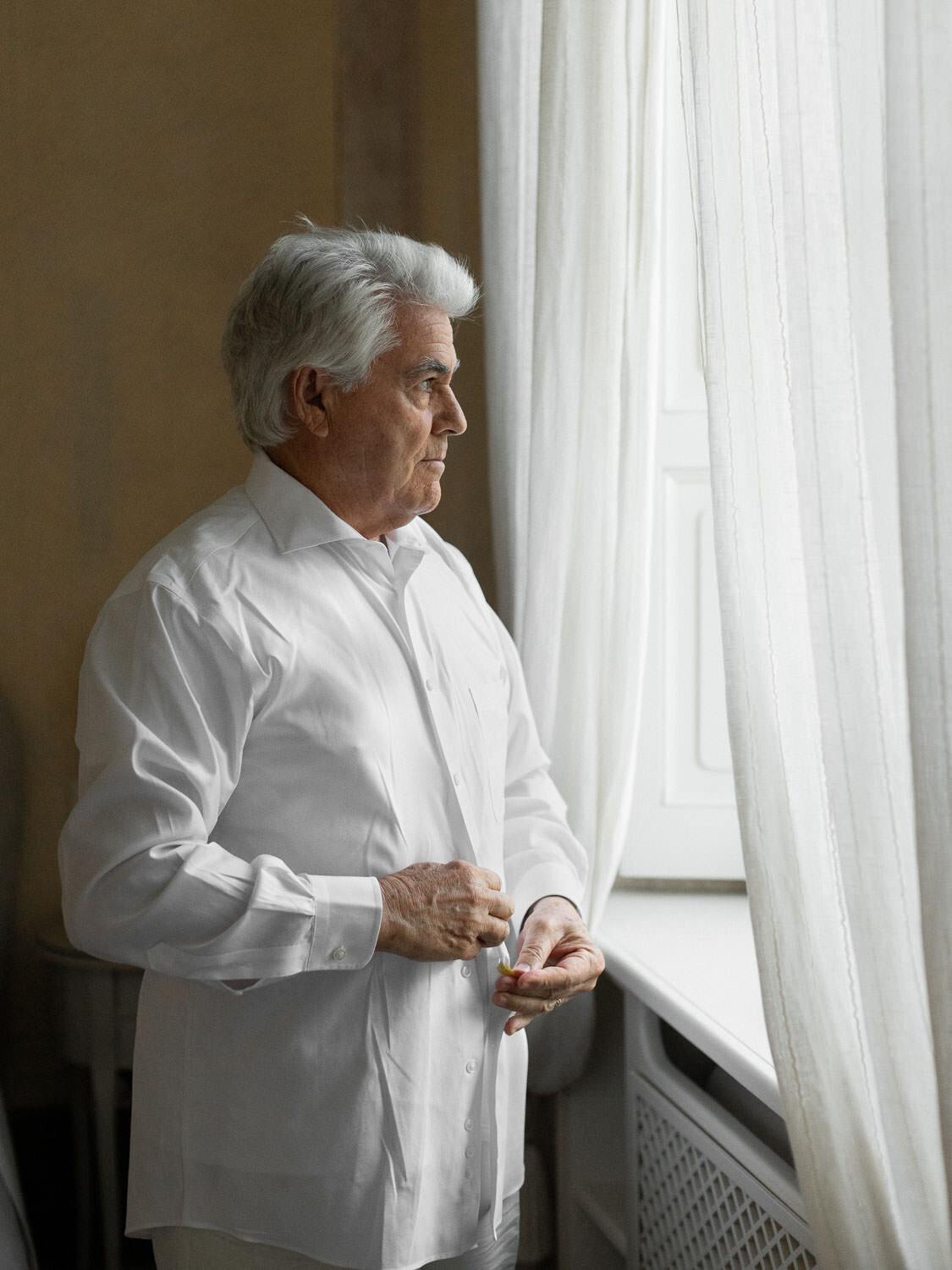 grey headed father buttoning white shirt in front of window