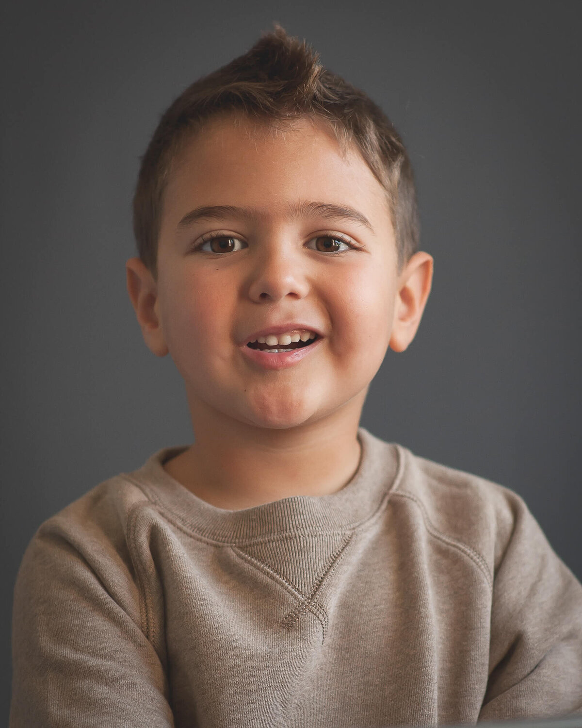 young boy wearing a brown sweatshirt making a silly face in a studio portrait with a grey background