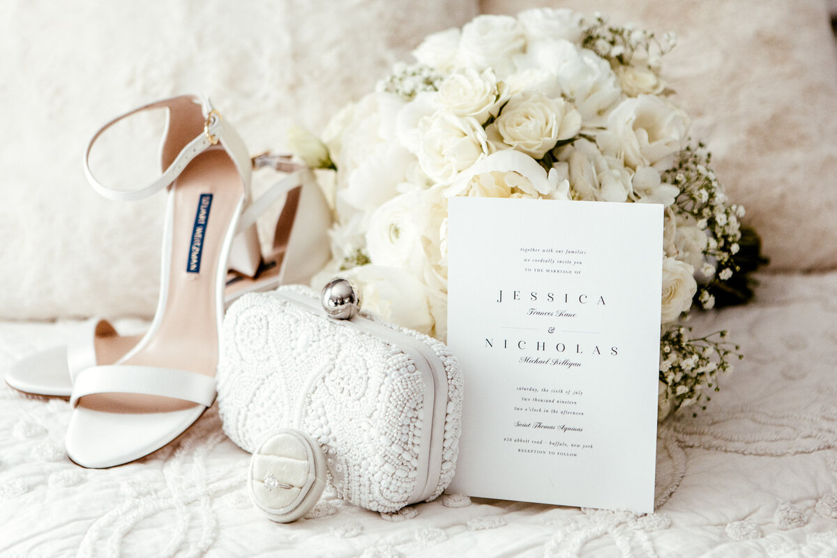 Wedding invitation against bouquet of white roses and among white heels, lace clutch bag and wedding ring