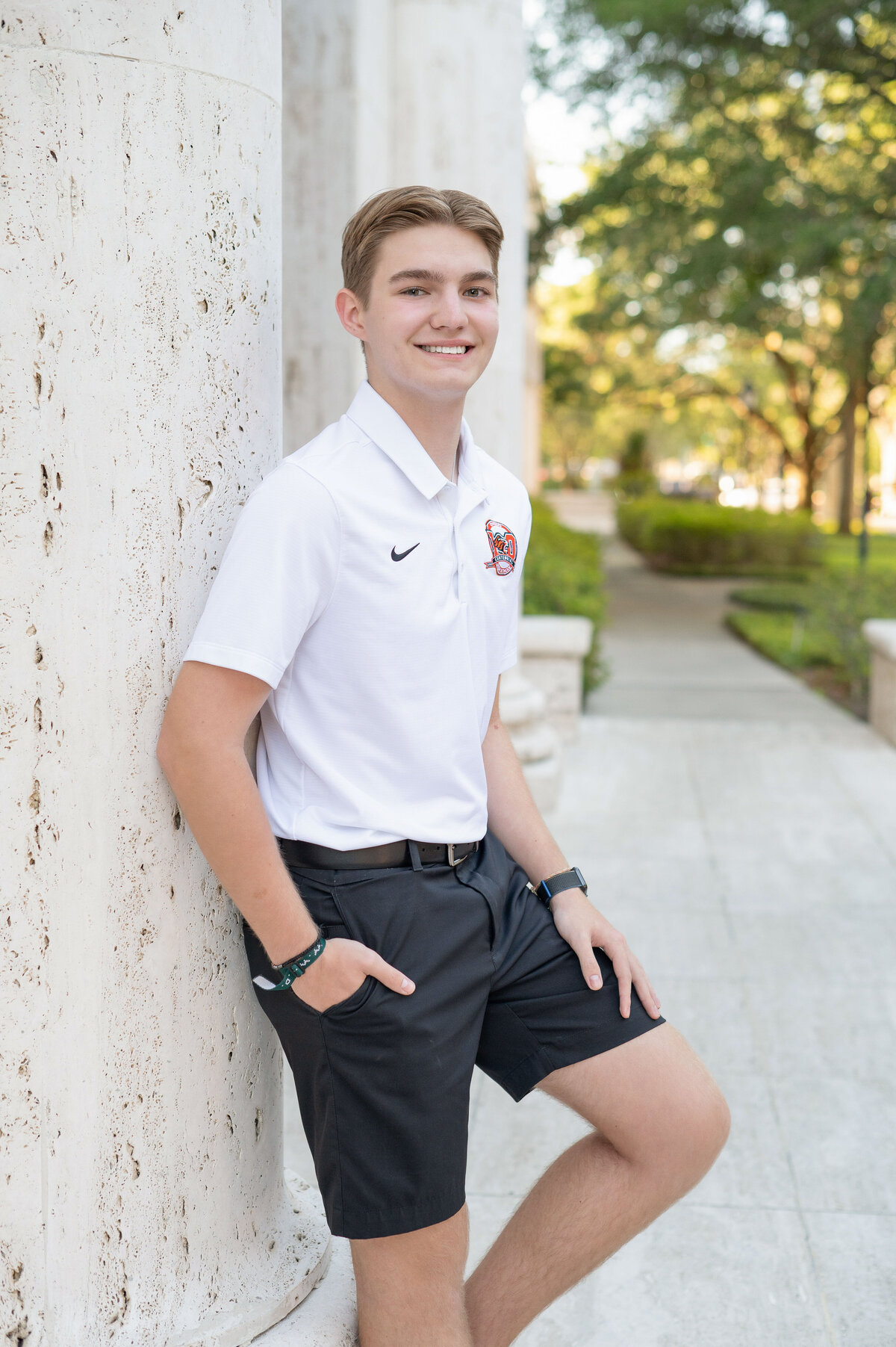 High school senior boy wearing a white polo leaning against a pillar.