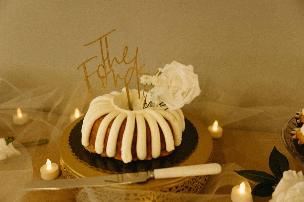 A decorative bundt cake with white icing and a "the forks" topper, displayed on a golden tray surrounded by lit candles and white flowers, perfect for Iowa weddings.
