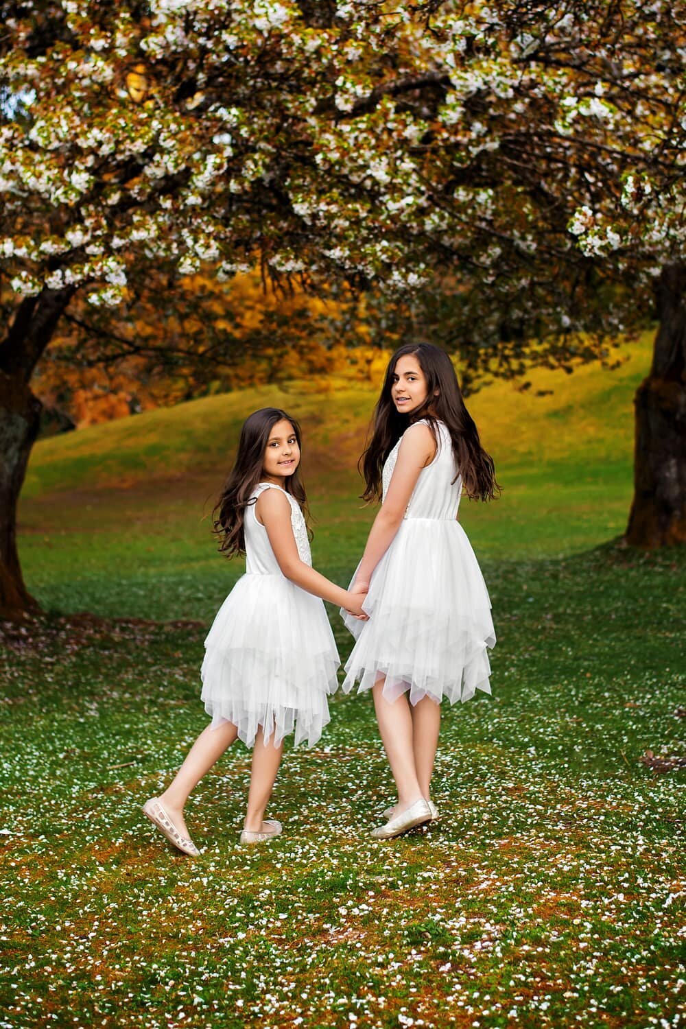 Sisters wearing white dresses holding hands in the cherry blossoms at Queen Elizabeth Park