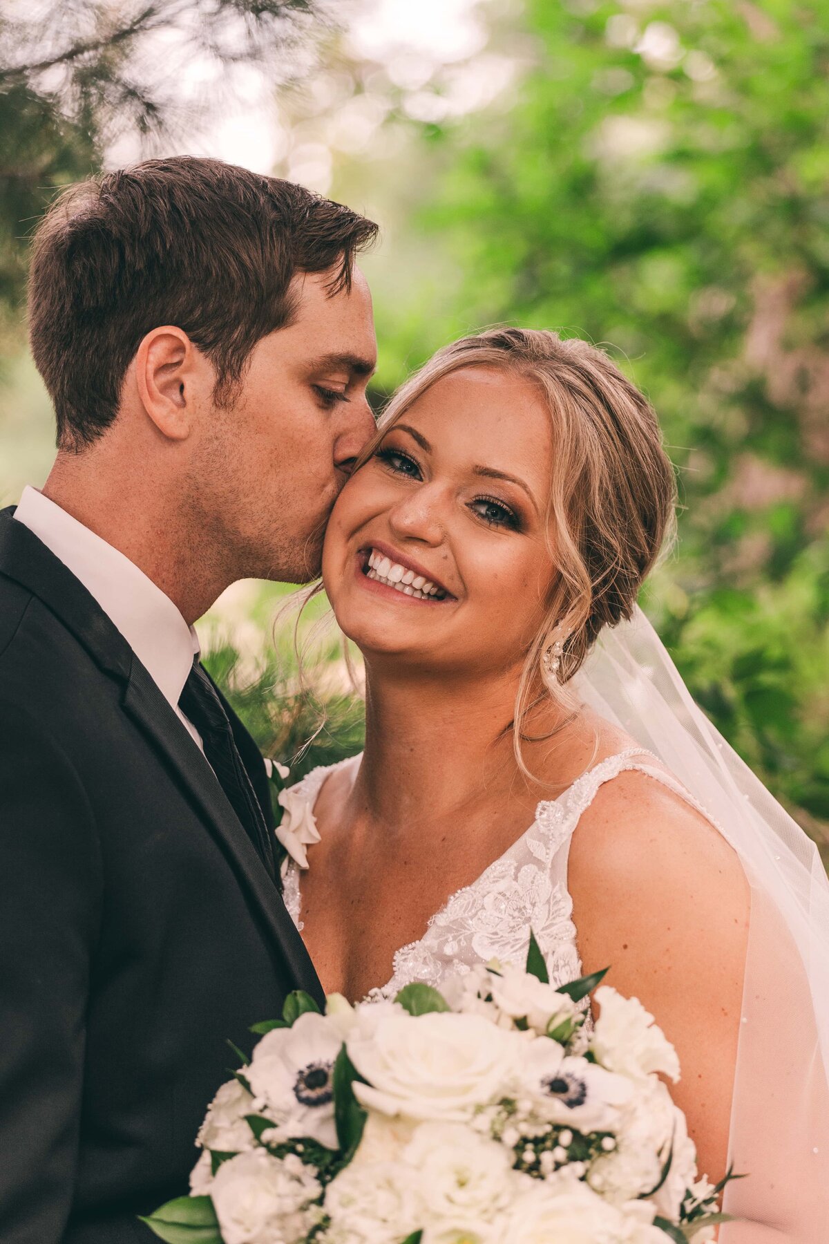 Groom kissing bride on the cheek as she smiles, both dressed in wedding attire, holding a bouquet, with an Iowa greenery background.