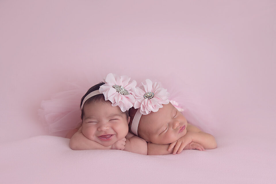 Smiling newborn baby girl twins lay on a pink bed wearing matching floral headbands during their NJ newborn photography session