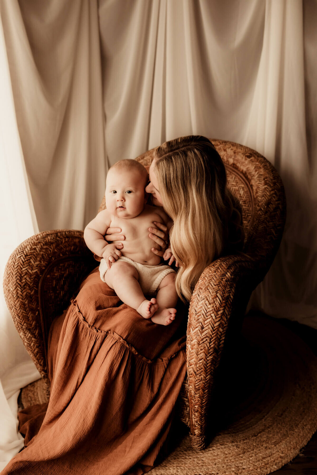 Milestone photo of a baby boy sitting in his mother's lap on a rattan chair with drapes behind them.